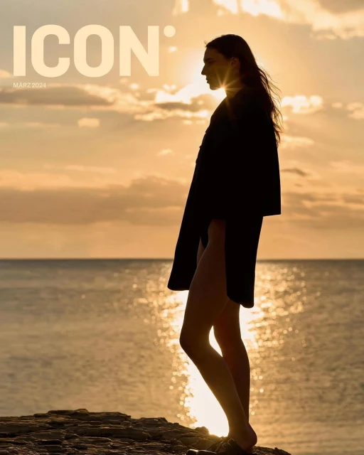 Silhouette of a woman in a long coat standing on a rocky outcrop by the sea at sunset, magazine cover for ICON featuring visible text.