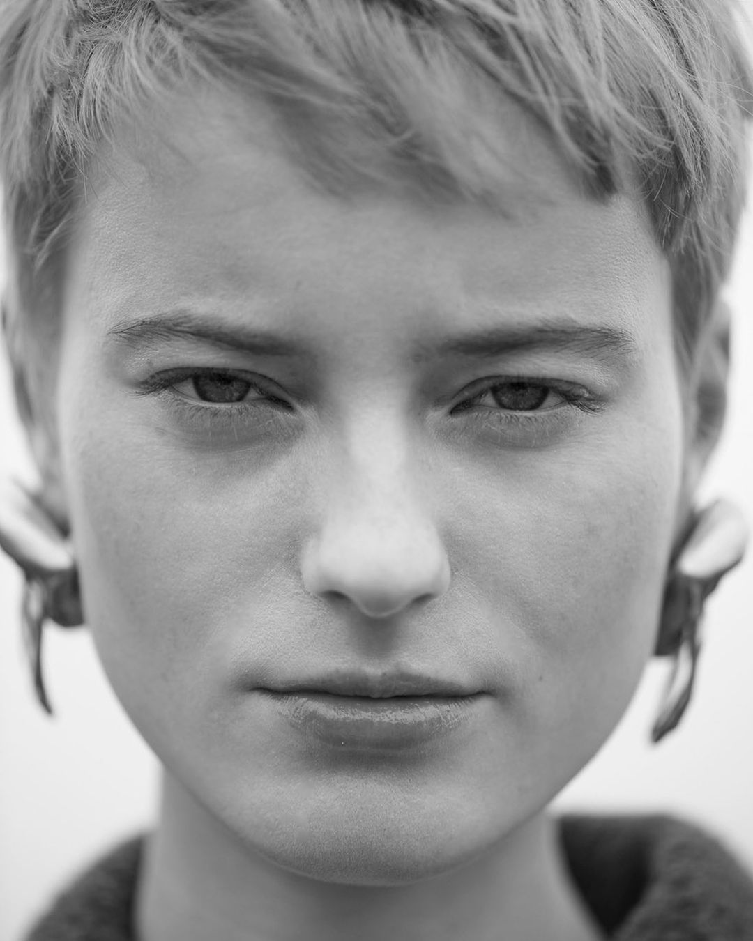 Monochrome portrait of a young adult with short hair, neutral expression, and statement earrings against a bright background.