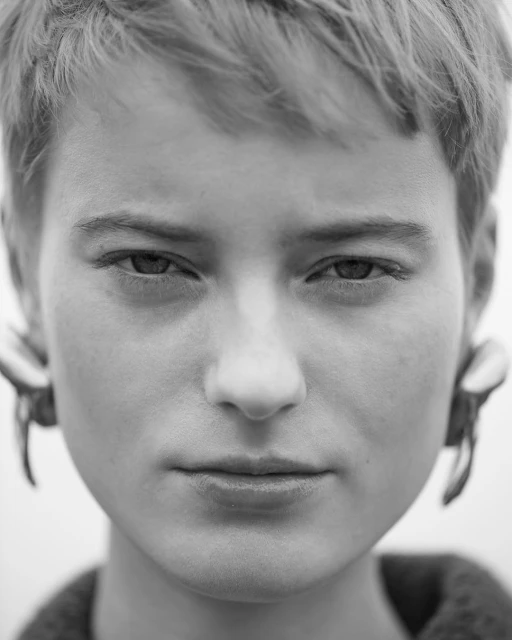Monochrome portrait of a young adult with short hair, neutral expression, and statement earrings against a bright background.