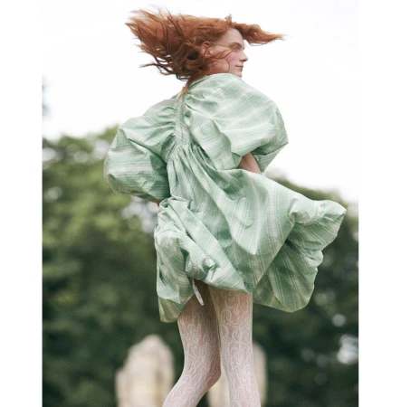 Model with red hair wearing a voluminous green dress and white patterned tights against an outdoor backdrop.