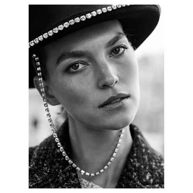 Monochrome portrait of a woman with a hat, wearing a rhinestone chain and necklace, with a serious expression, shot in medium close-up.