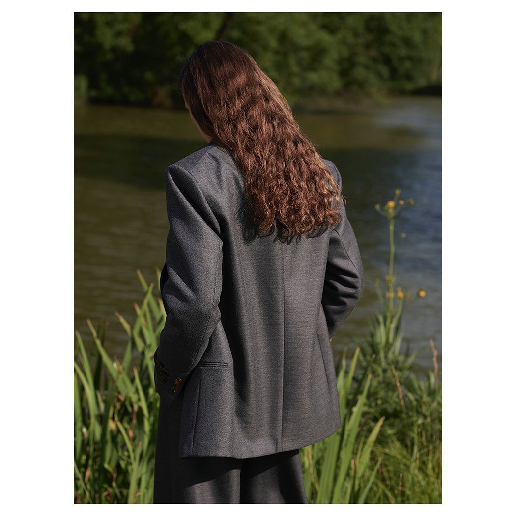 Back view of an adult woman with long curly auburn hair wearing a gray suit outdoors, with a body of water and greenery in the background.
