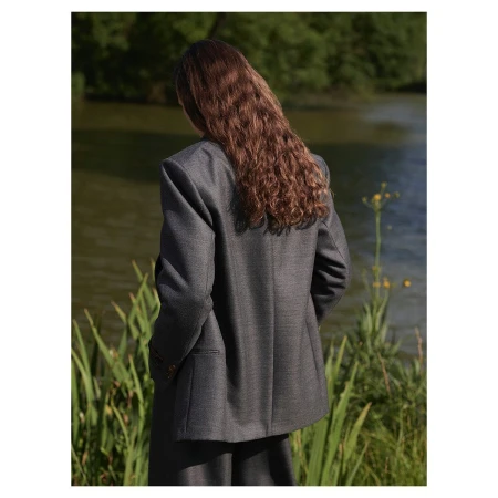 Woman with long wavy brown hair wearing a grey blazer standing outdoors near water and tall reeds