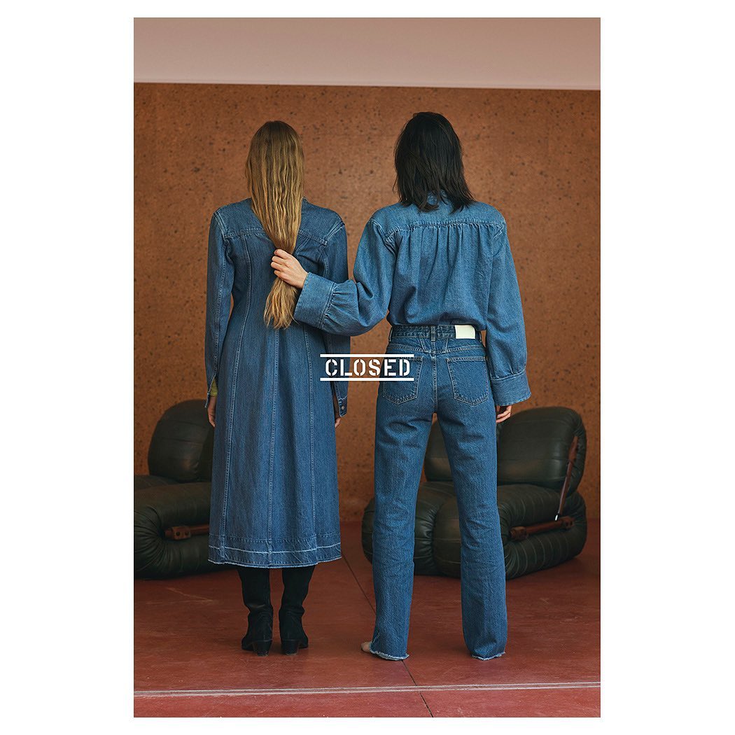 Two women in matching denim outfits pose indoors against a textured wall with an armchair. One touches the other’s long blonde hair.