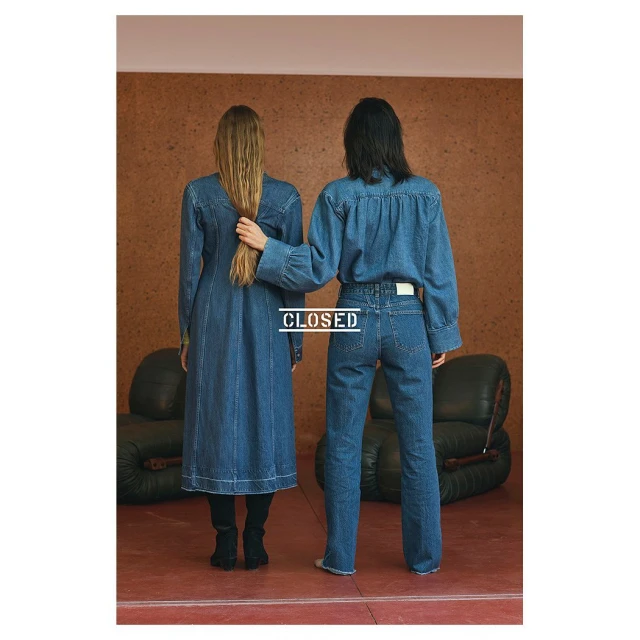 Two women in matching denim outfits pose indoors against a textured wall with an armchair. One touches the other’s long blonde hair.