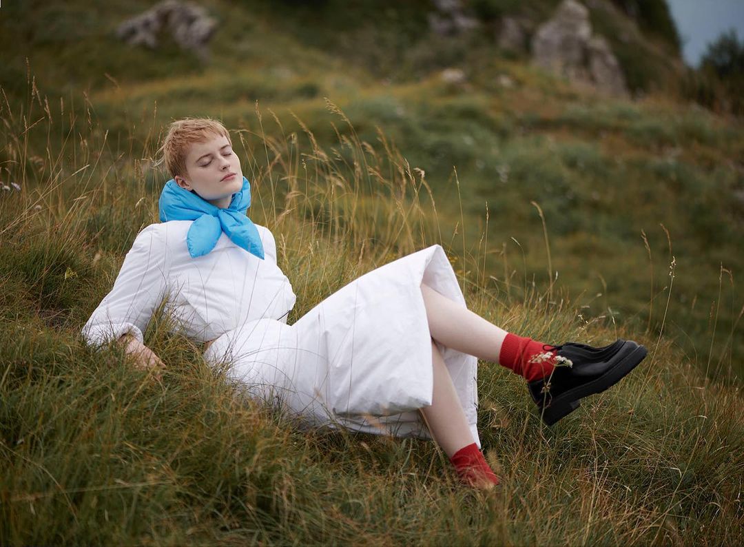 An adult female with short hair lies on a grassy hill wearing a white dress and blue scarf, eyes closed in a peaceful outdoor setting.