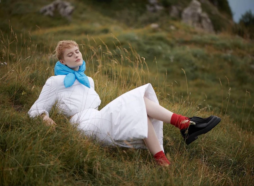 An adult female with short hair lies on a grassy hill wearing a white dress and blue scarf, eyes closed in a peaceful outdoor setting.