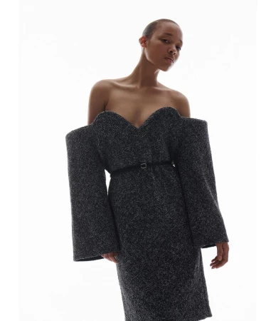 Model with a shaved head wearing a dark textured off-the-shoulder dress against a bright white background