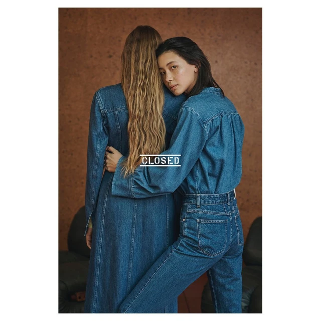 Two women in denim outfits embrace in a fashion studio setting. One woman faces the camera, showcasing the brand's denim collection.