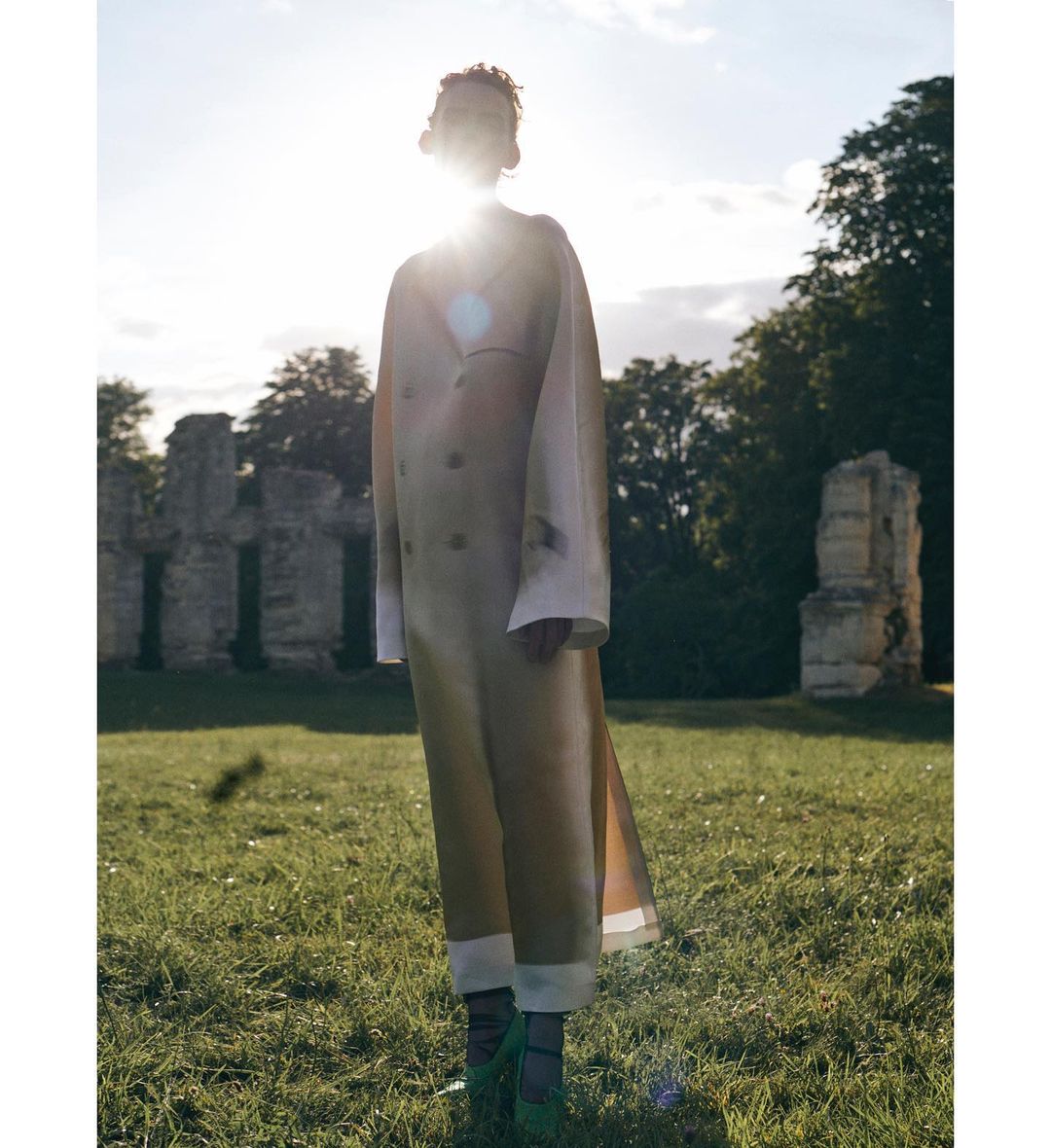 A single woman standing in a field wearing a long beige coat, green shoes, and black tights, with ruined walls and green trees in the background.