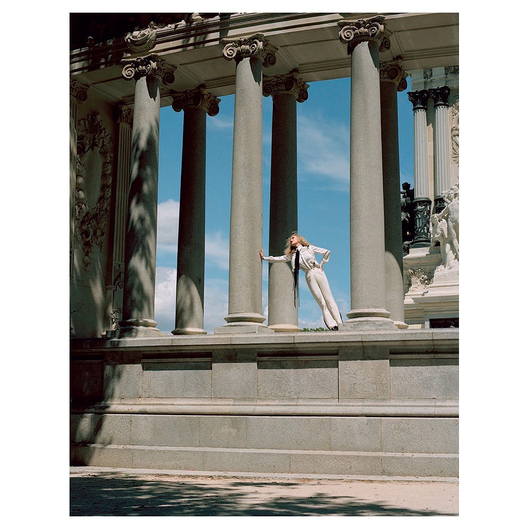 Adult female model in white suit poses between stone columns against a blue sky in an outdoor architectural setting. Fashion shoot.