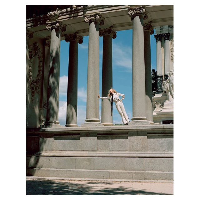Adult female model in white suit poses between stone columns against a blue sky in an outdoor architectural setting. Fashion shoot.