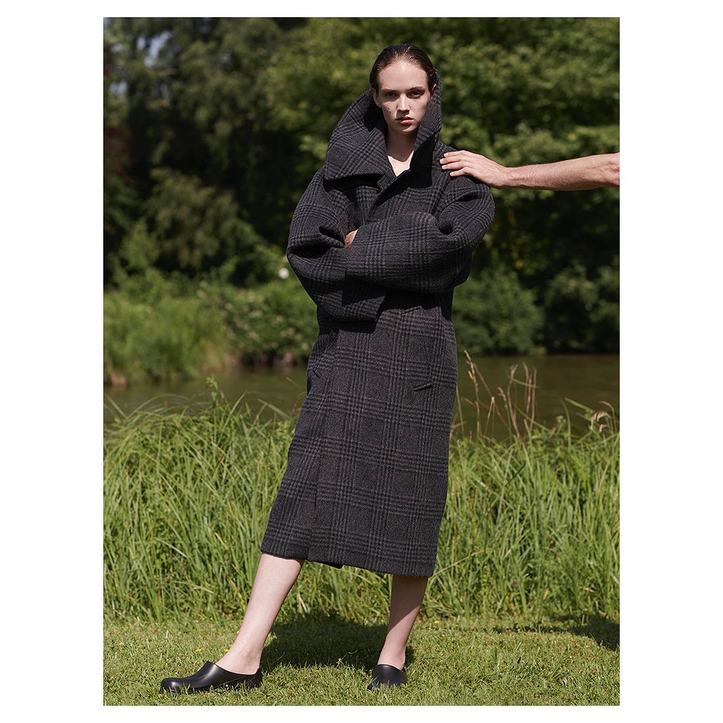 Fashion shot of a woman wearing a gray plaid coat and skirt with an arm resting on her shoulder in a grassy outdoor setting.