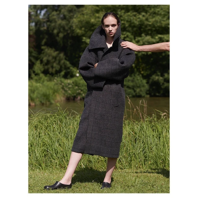 Fashion shot of a woman wearing a gray plaid coat and skirt with an arm resting on her shoulder in a grassy outdoor setting.