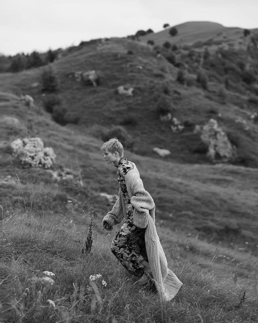 A young adult woman in a patterned outfit and long cardigan is walking up a grassy hill in a mountainous, black and white outdoor setting.