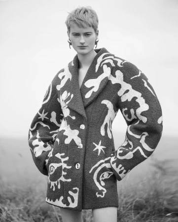 Model with short hair wearing a patterned coat with sculptural shoulders against an outdoor landscape background