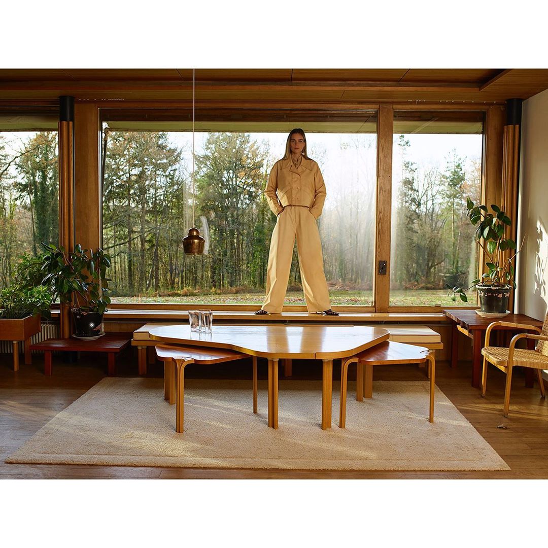 A woman in a beige jumpsuit poses in front of a window in a modern interior with light wood, looking at the camera with a neutral expression.
