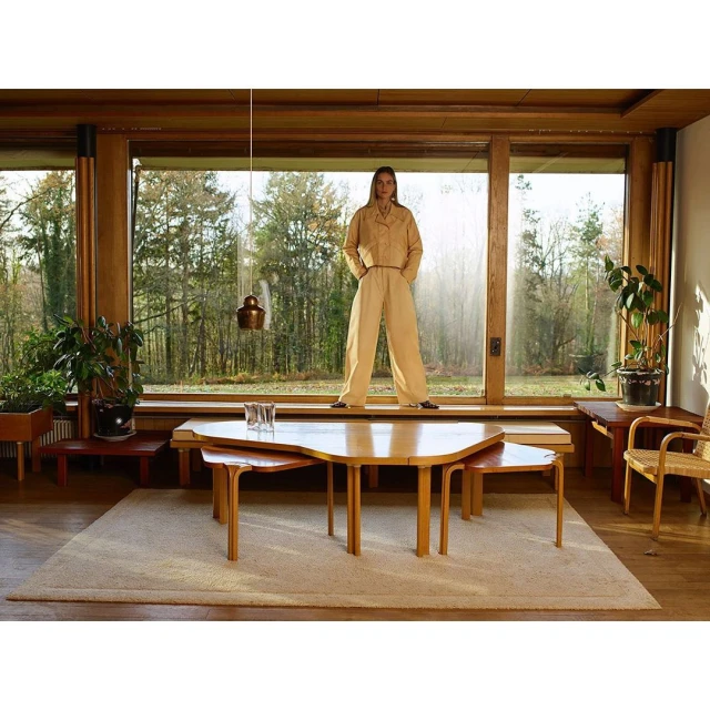 A woman in a beige jumpsuit poses in front of a window in a modern interior with light wood, looking at the camera with a neutral expression.