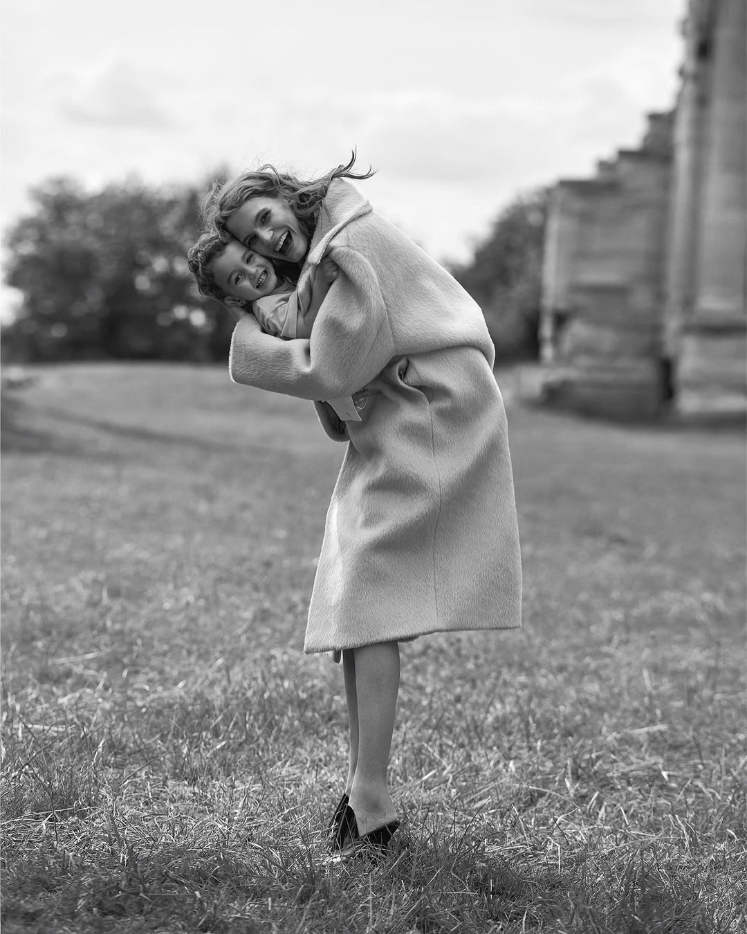 Black and white portrait of a young woman in a coat carrying a child in a grassy field with architectural ruins in the background.