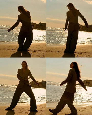 A person in a blazer and wide-legged trousers posing on a beach, the warm light creating a silhouette effect as they walk in the sand.