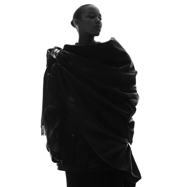 Black and white portrait of a slender woman wearing a black draped garment against a bright white background in a studio setting.