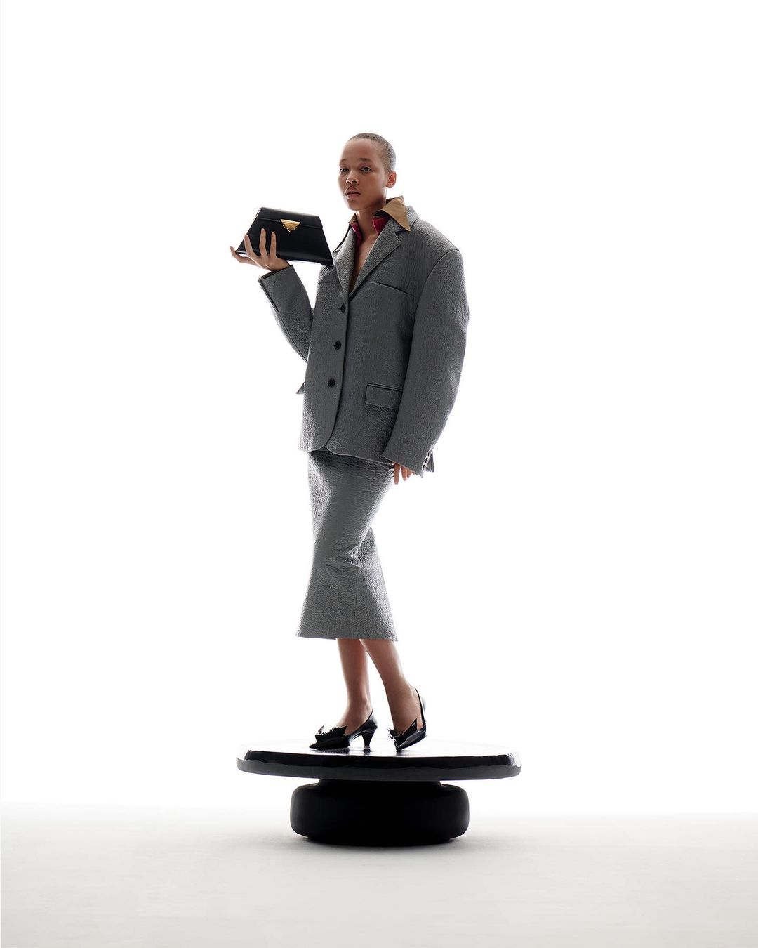 A woman in a gray suit and black high heels stands on a black platform while holding a black Prada clutch against a white background.