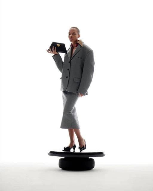 A woman in a gray suit and black high heels stands on a black platform while holding a black Prada clutch against a white background.