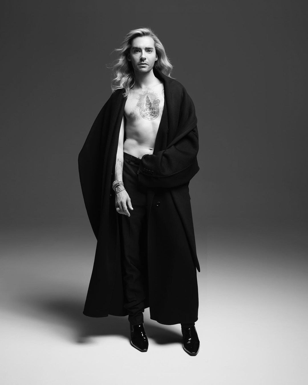 Black and white portrait of a blonde male model with tattoo, wearing a black robe and patent leather boots, posing in a studio.