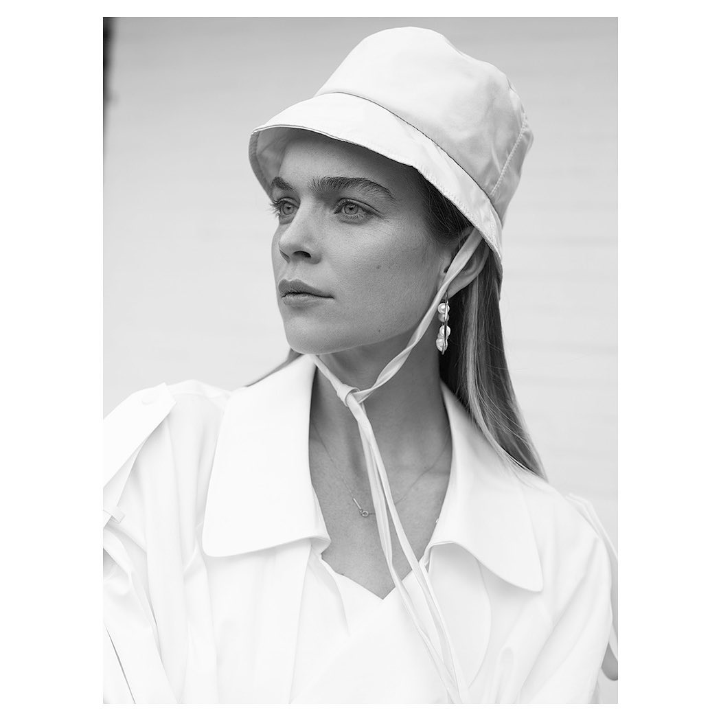 Black and white portrait of a young woman wearing a white bucket hat and a white jacket with statement earrings looking off-camera.