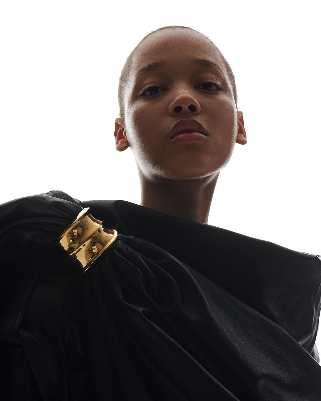Low-angle portrait of a young woman with short hair wearing a draped black dress with a gold clasp against a white background.