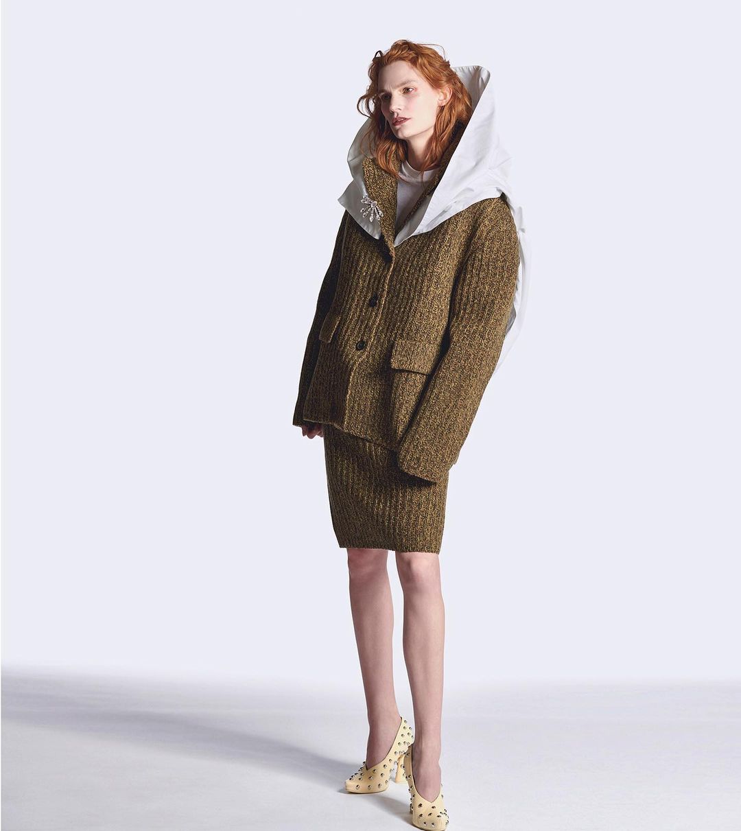 Red-haired model in a textured brown jacket and skirt with embellished cream heels, posing against a gradient white backdrop.