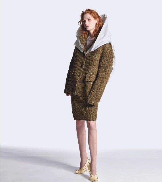 Red-haired model in a textured brown jacket and skirt with embellished cream heels, posing against a gradient white backdrop.