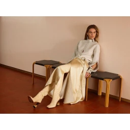 Model wearing a light grey jacket and long satin skirt seated on black and wooden benches against a pink wall.