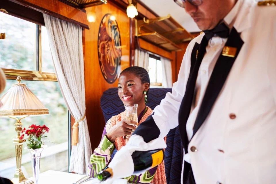 Woman smiles while being served champagne by a server in formal wear, inside a train car with table lamp and flower arrangement.