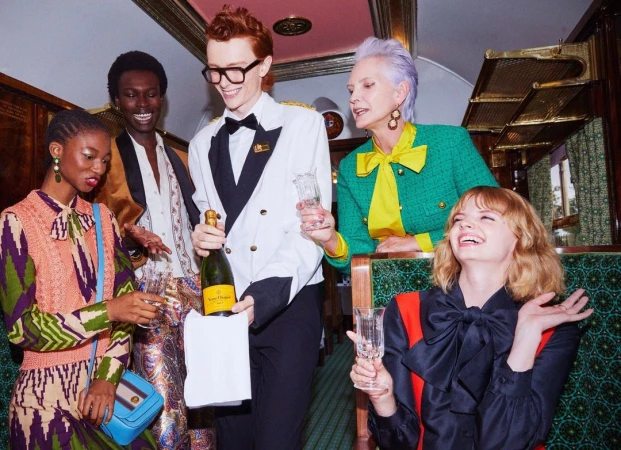 Five people celebrate with champagne inside a train car. The group includes men and women in formal wear, seated and standing.