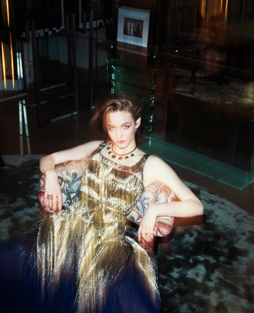 Young woman in a gold gown sitting in patterned armchair inside. She wears a statement necklace and several rings. Dark mood lighting.
