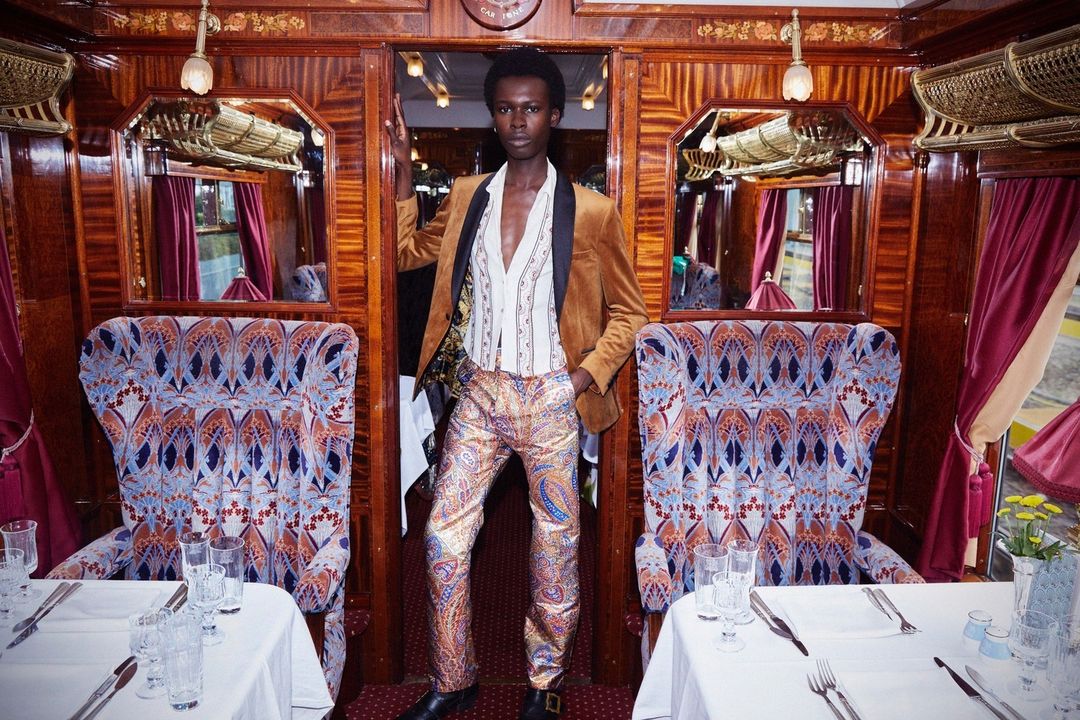 A man in a brown jacket and paisley pants stands in a vintage train car with set dining table, showcasing high fashion in an opulent interior.