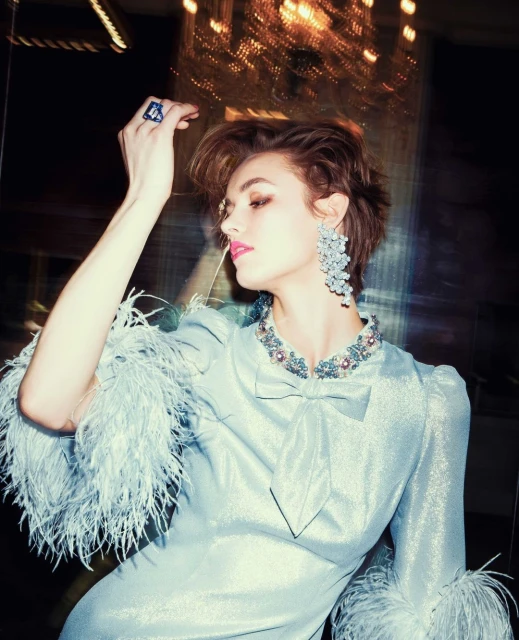 Young woman in a light blue dress with feathered sleeves and statement jewelry poses indoors under warm lighting with blurred effects.