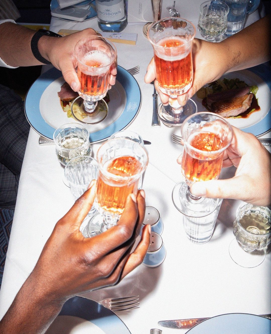 Overhead shot of four people toasting with glasses of a pink drink at a table set with plates of food, cutlery, and a water bottle.