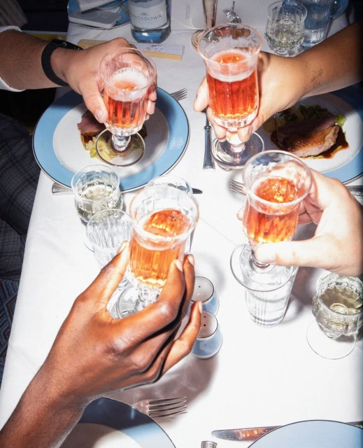 Overhead shot of four people toasting with glasses of a pink drink at a table set with plates of food, cutlery, and a water bottle.