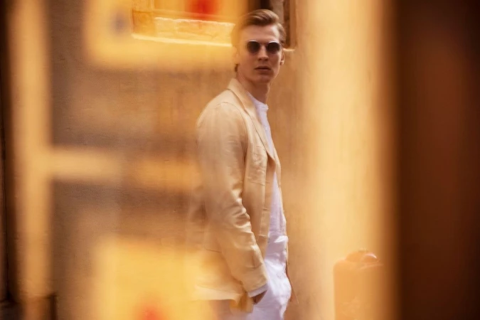 Man wearing round sunglasses and light beige blazer standing outdoors with foreground obstruction