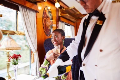 A smiling black woman holding a champagne flute inside a luxury train carriage as a steward pours champagne.