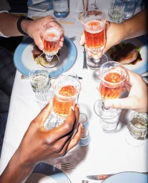 Four hands holding small ornate crystal glasses filled with amber liquid in a toast over a white tablecloth dinner setting.