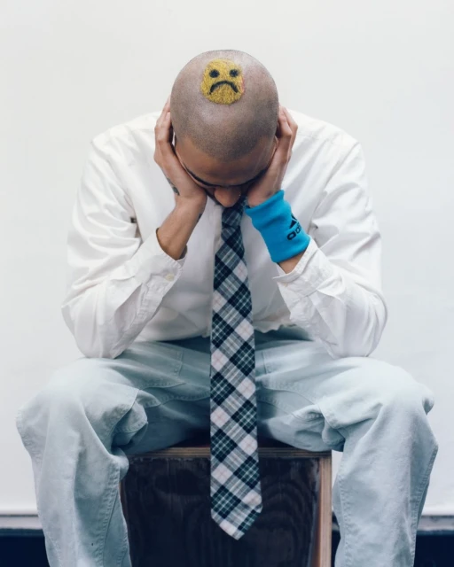Person with shaved head sitting, wearing a tie and a sad-face sticker on head, head in hands, wristband on wrist, indoors.