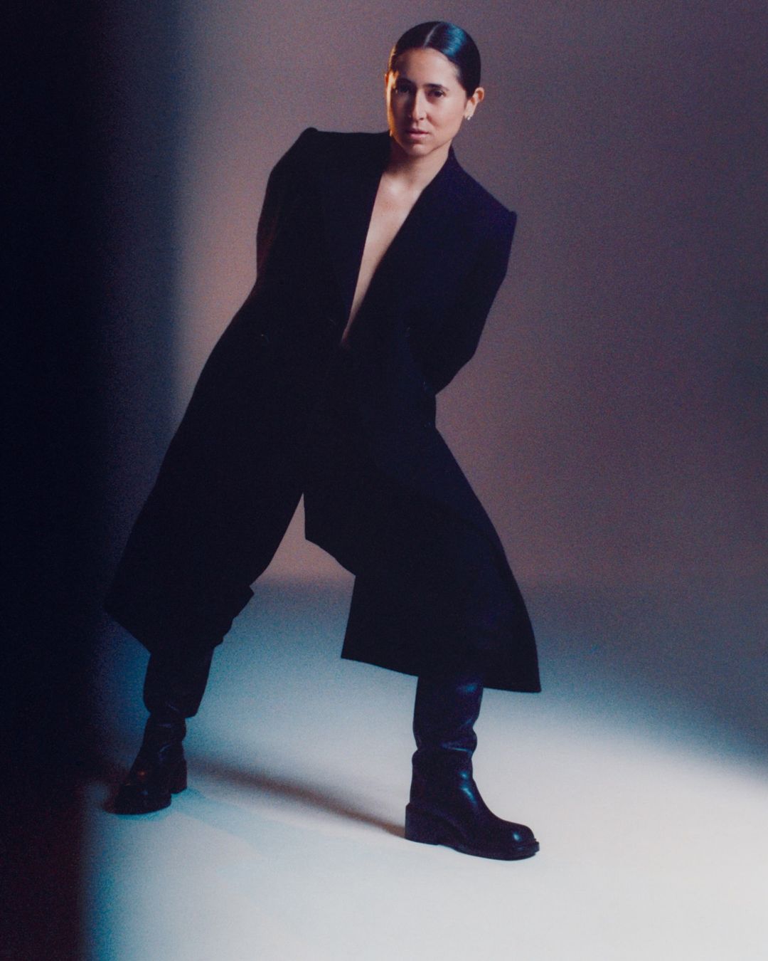 Woman in dark coat and boots posing against a studio backdrop under moody lighting