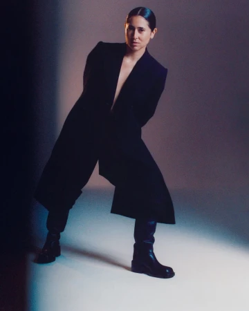 Woman in dark coat and boots posing against a studio backdrop under moody lighting