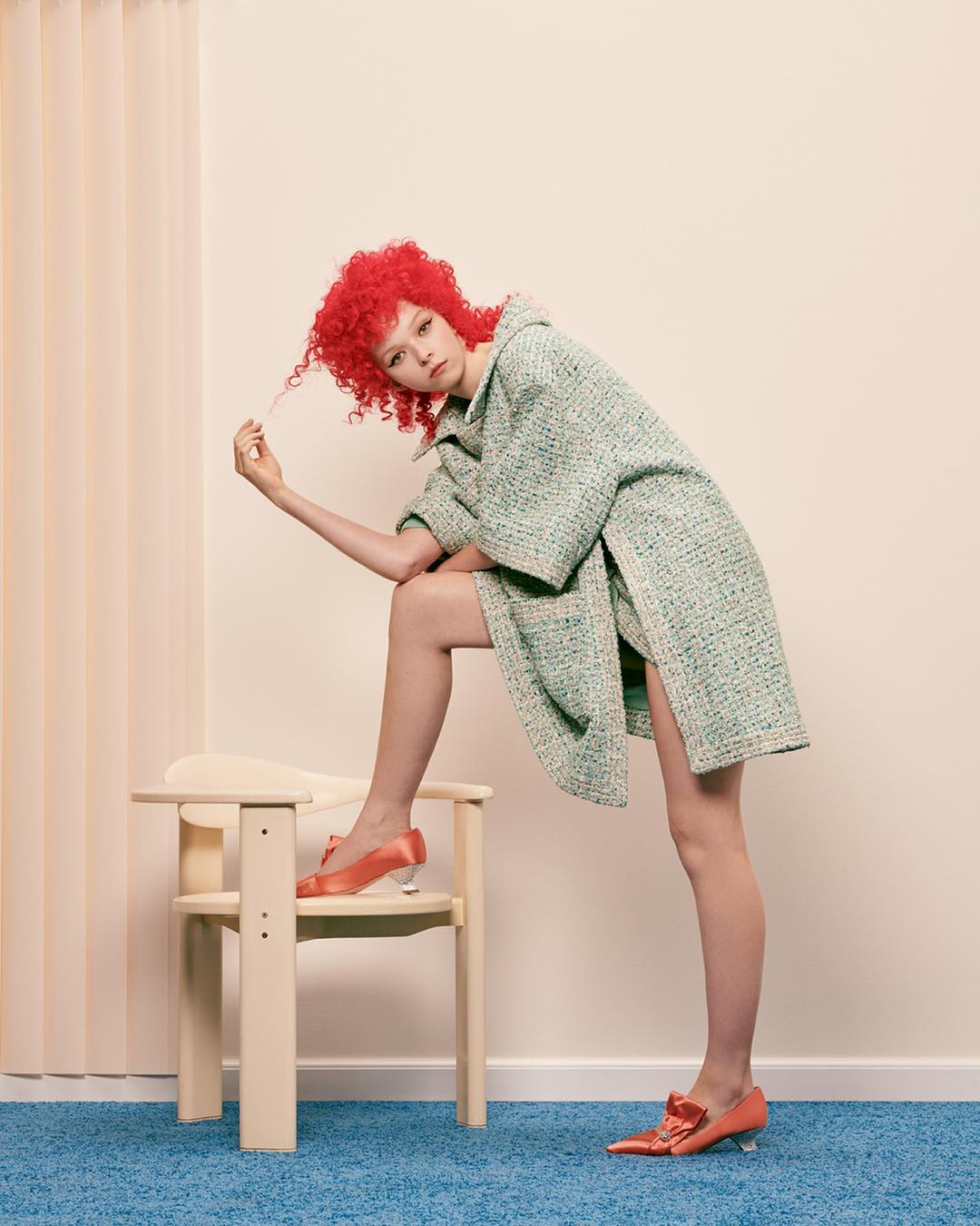 Fashion shot of a stylish woman with curly red hair, wearing a green tweed coat, and salmon heels. She is posing with a chair.