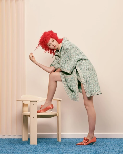Fashion shot of a stylish woman with curly red hair, wearing a green tweed coat, and salmon heels. She is posing with a chair.
