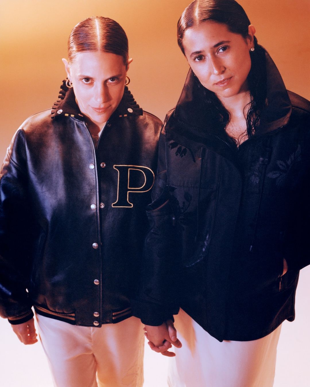 Two women stand in a close-up shot, wearing jackets and light pants, holding hands against a peach-colored backdrop in a studio setting.