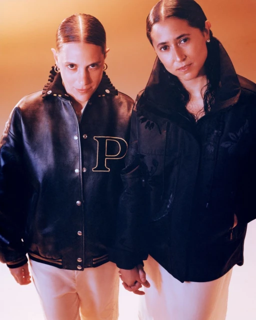 Two women stand in a close-up shot, wearing jackets and light pants, holding hands against a peach-colored backdrop in a studio setting.
